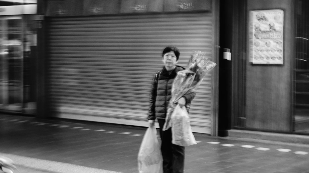 black and white of dreary middle aged woman holding two bouquets of flowers and a bag of groceries 